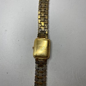 May include: A gold-toned wristwatch with a rectangular face and a linked band. The watch face has a gold finish, and the band is made of interlocking, textured links. The back of the watch has text that is difficult to read.