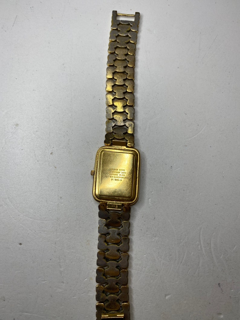 May include: A gold-toned wristwatch with a rectangular face and a linked band. The watch face has a gold finish, and the band is made of interlocking, textured links. The back of the watch has text that is difficult to read.