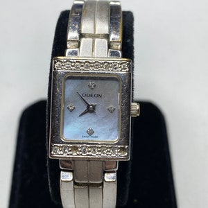 K&ouml;nnte beinhalten: Silberfarbene Uhr mit quadratischem Zifferblatt und Perlmutt-Zifferblatt. Die Uhr hat ein silberfarbenes Armband mit einem diamantbesetzten L&uuml;nette. Das Zifferblatt der Uhr tr&auml;gt die Aufschriften "ODEON" und "SWISS MADE".