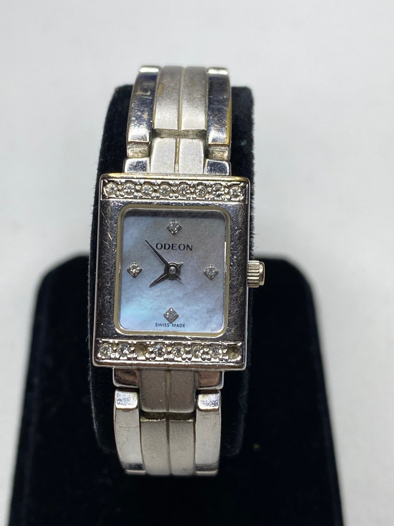 K&ouml;nnte beinhalten: Silberfarbene Uhr mit quadratischem Zifferblatt und Perlmutt-Zifferblatt. Die Uhr hat ein silberfarbenes Armband mit einem diamantbesetzten L&uuml;nette. Das Zifferblatt der Uhr tr&auml;gt die Aufschriften "ODEON" und "SWISS MADE".