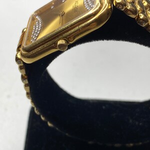 May include: A gold-toned Heloisa Swiss wristwatch with a rectangular face. The watch features a gold band and a gold face with diamond accents. The watch is displayed on a black velvet surface.
