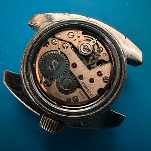 May include: Close-up of the back of a silver wristwatch with a round, clear case revealing the inner workings of the watch. The watch face has the text "SWISS 17 JEWELS" printed on it.