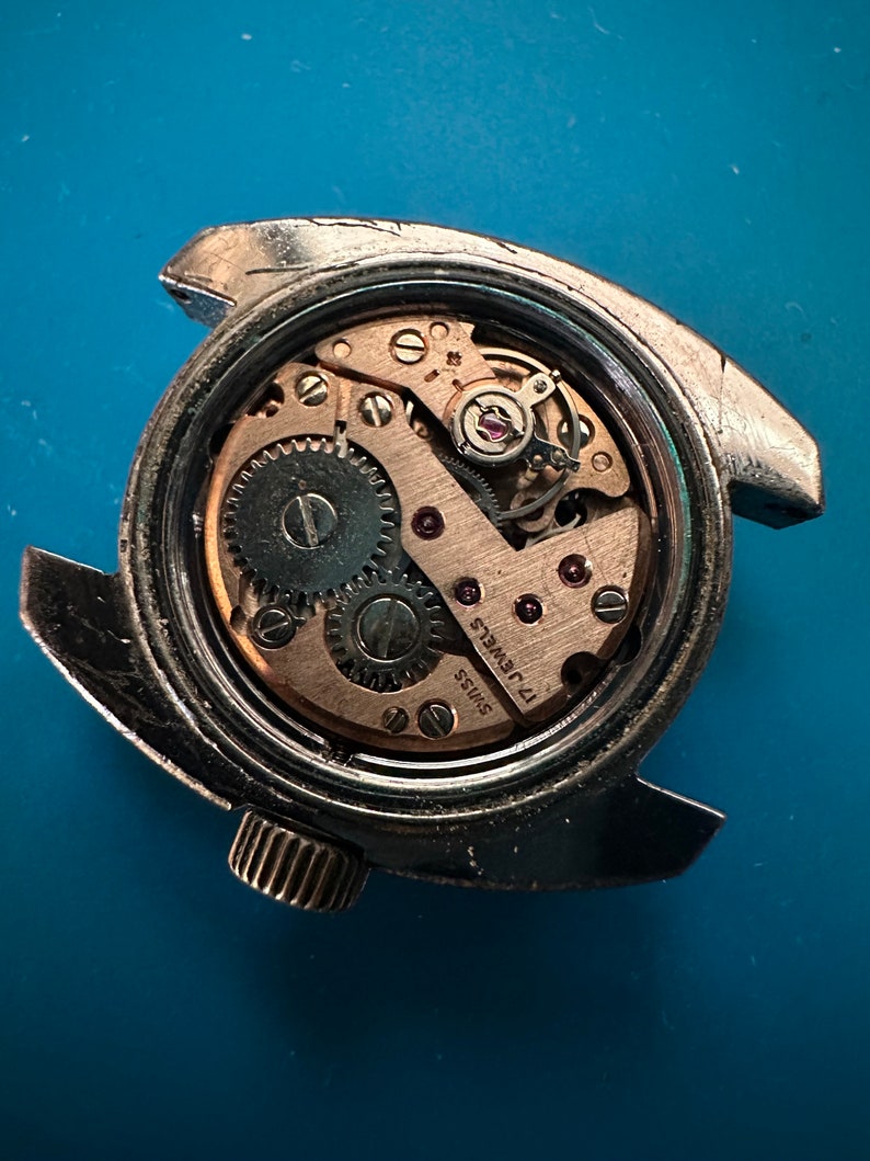 May include: Close-up of the back of a silver wristwatch with a round, clear case revealing the inner workings of the watch. The watch face has the text "SWISS 17 JEWELS" printed on it.