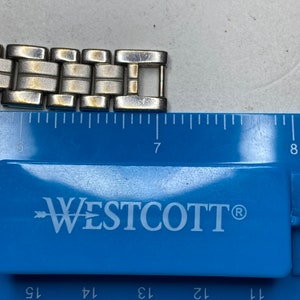 K&ouml;nnte beinhalten: Ein silbernes und goldenes Armband mit einem Verschluss. Das Armband wird an einem blauen Lineal mit dem Markennamen Westcott gemessen.