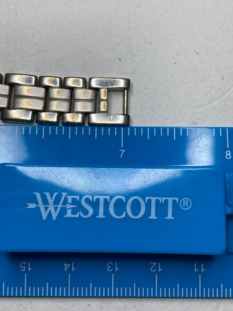 K&ouml;nnte beinhalten: Ein silbernes und goldenes Armband mit einem Verschluss. Das Armband wird an einem blauen Lineal mit dem Markennamen Westcott gemessen.