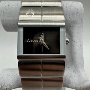 May include: A silver Nixon watch with a black face and a stainless steel band. The watch has a square face and a simple design.