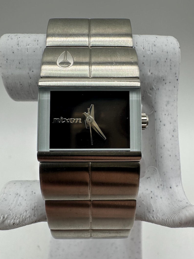 May include: A silver Nixon watch with a black face and a stainless steel band. The watch has a square face and a simple design.