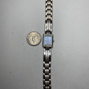 K&ouml;nnte beinhalten: Eine silberne Uhr mit quadratischem Zifferblatt und einem silbernen Metallband. Das Zifferblatt hat einen blauen Hintergrund mit silbernen Zeigern und einer silbernen L&uuml;nette mit kleinen, klaren Steinen.