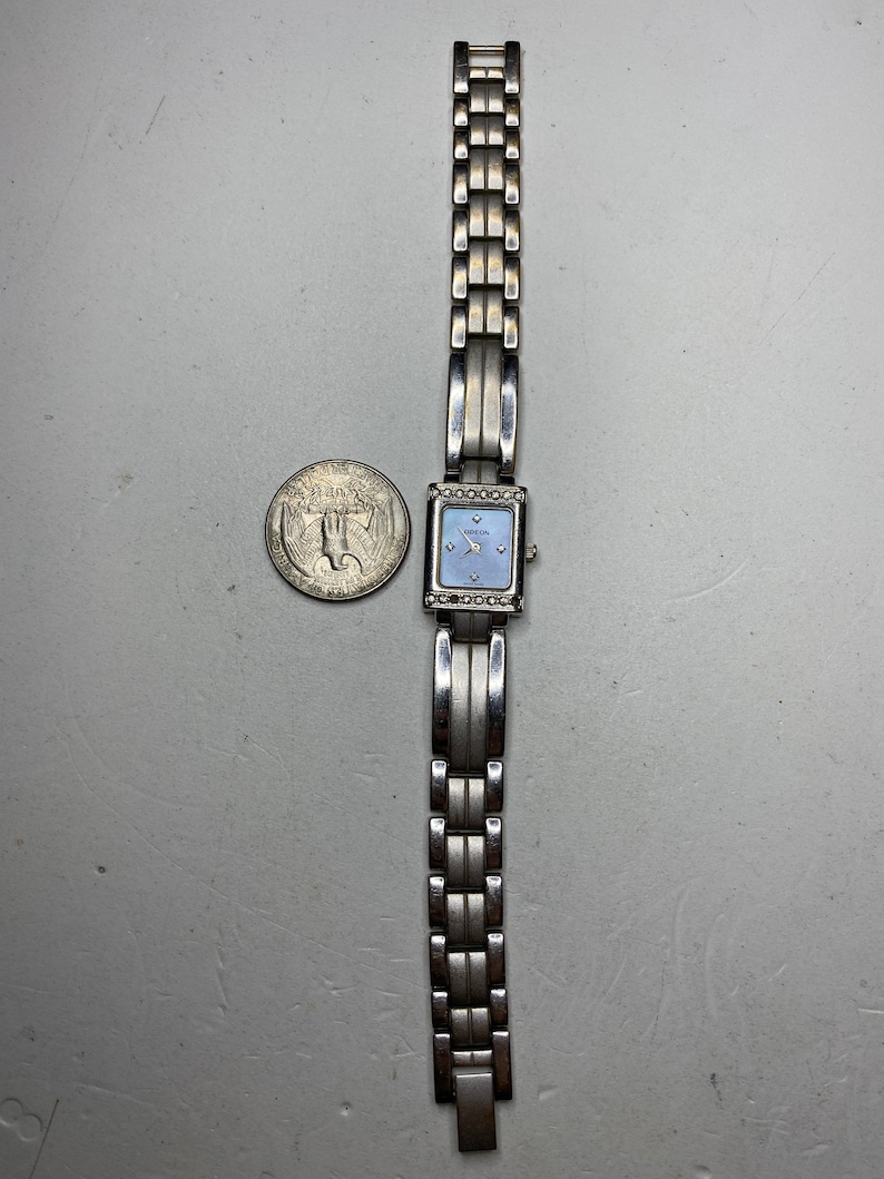 K&ouml;nnte beinhalten: Eine silberne Uhr mit quadratischem Zifferblatt und einem silbernen Metallband. Das Zifferblatt hat einen blauen Hintergrund mit silbernen Zeigern und einer silbernen L&uuml;nette mit kleinen, klaren Steinen.