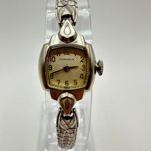 May include: A silver wristwatch with a square face and a silver metal band. The watch face has black Roman numeral hour markers and a white dial with the word "Caravelle" printed on it. The watch is made in Switzerland.