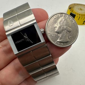 May include: A silver Nixon watch with a black face and a silver band. The watch is about the size of a US quarter.