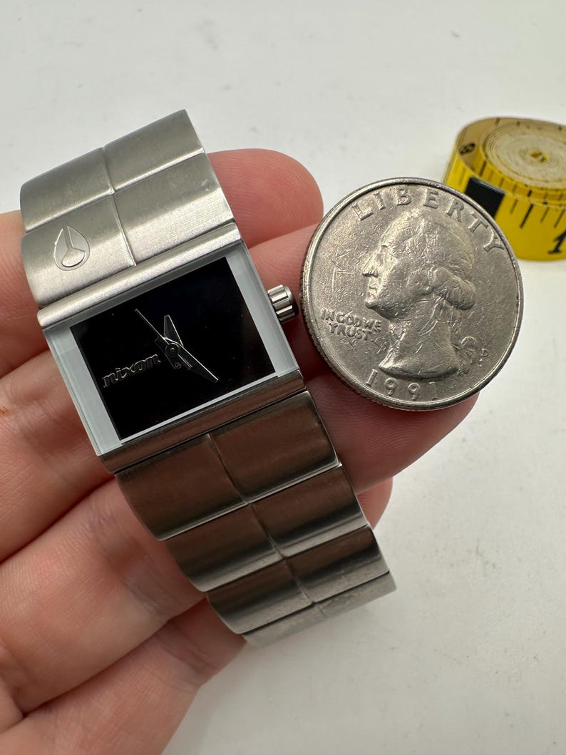 May include: A silver Nixon watch with a black face and a silver band. The watch is about the size of a US quarter.
