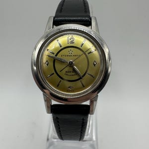 May include: A silver wristwatch with a black leather band. The watch face is yellow with black markings and the text "ETERNA-MATIC" and "BIRK" "CHALLENGER" in black. The watch face also has a diamond-shaped marker at the 12 o'clock position.