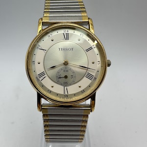 May include: A gold and silver two-tone Tissot wristwatch with a white face and black Roman numeral hour markers. The watch has a small subdial at the 12 o'clock position and the text "TISSOT" printed on the face.