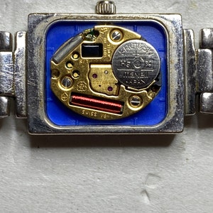 K&ouml;nnte beinhalten: Nahaufnahme der R&uuml;ckseite einer silbernen Uhr mit quadratischem Zifferblatt. Die Uhr hat eine blaue R&uuml;ckseite und ein silbernes Armband. Das Zifferblatt der Uhr hat einen silbernen und goldenen Mechanismus mit einer runden silbernen Batterie mit der Aufschrift "SR616SW" und "MENEB JAPAN".