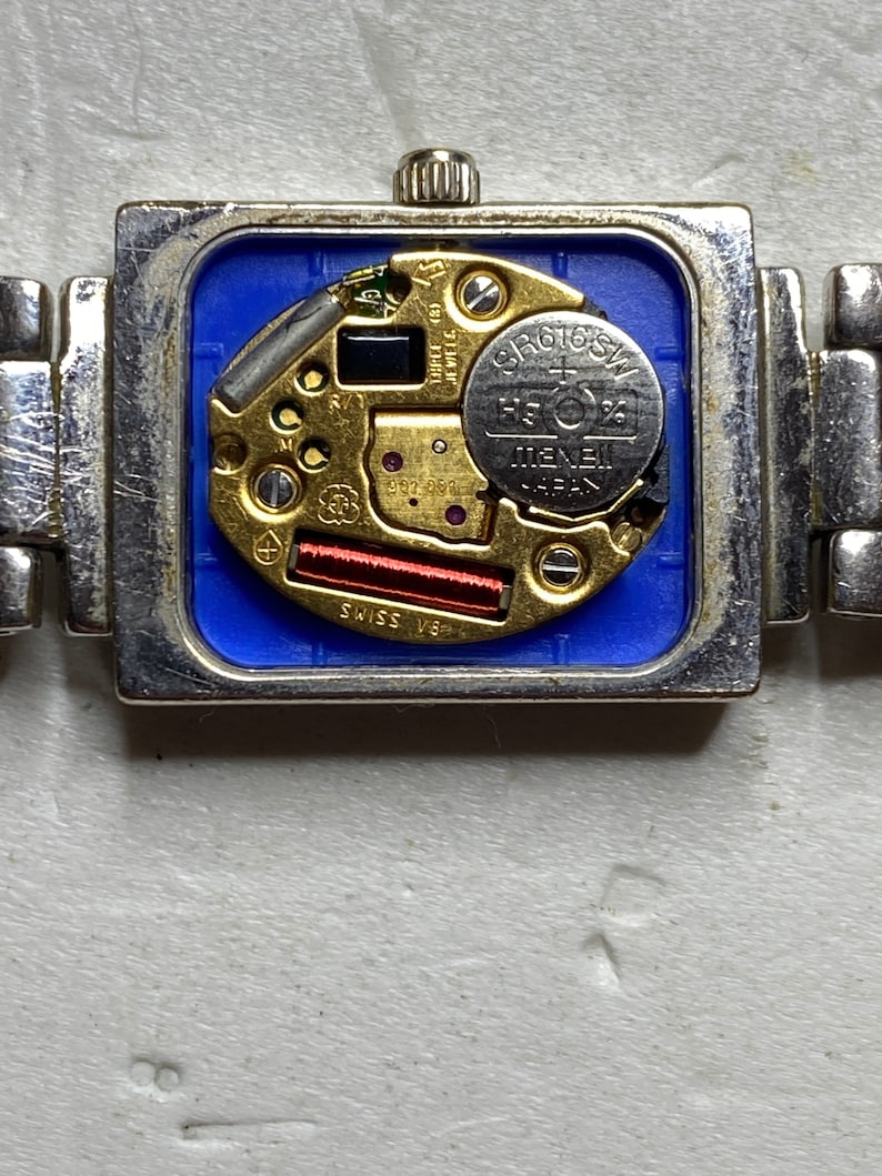 K&ouml;nnte beinhalten: Nahaufnahme der R&uuml;ckseite einer silbernen Uhr mit quadratischem Zifferblatt. Die Uhr hat eine blaue R&uuml;ckseite und ein silbernes Armband. Das Zifferblatt der Uhr hat einen silbernen und goldenen Mechanismus mit einer runden silbernen Batterie mit der Aufschrift "SR616SW" und "MENEB JAPAN".