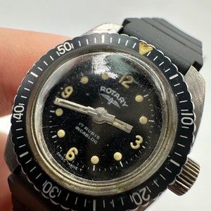 May include: A black Rotary wristwatch with a black face and white markings. The watch has a rotating bezel with white markings and a black rubber strap.