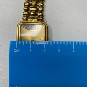 May include: A gold-toned wristwatch with a rectangular face and a linked band. The watch face features a gold dial with small, sparkling accents and the brand name "HELOISA SWISS". The band is made of rounded, gold-colored links.