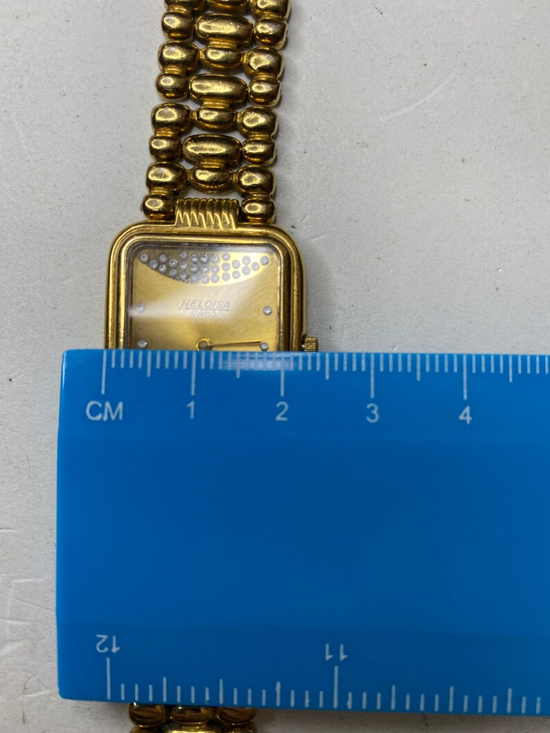 May include: A gold-toned wristwatch with a rectangular face and a linked band. The watch face features a gold dial with small, sparkling accents and the brand name "HELOISA SWISS". The band is made of rounded, gold-colored links.