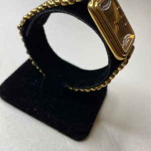 May include: A gold-toned wristwatch with a rectangular face and a textured band. The watch face is a warm brown color, with small diamond-like accents. The band is made of small, connected, rounded links.