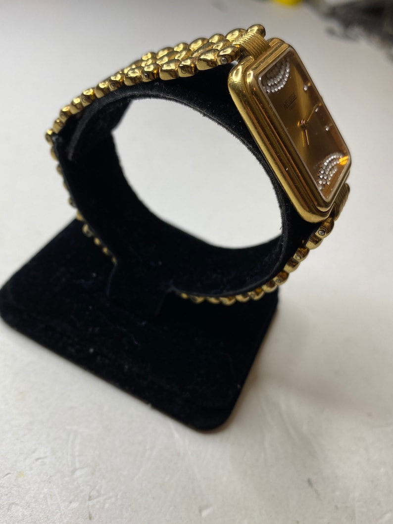 May include: A gold-toned wristwatch with a rectangular face and a textured band. The watch face is a warm brown color, with small diamond-like accents. The band is made of small, connected, rounded links.