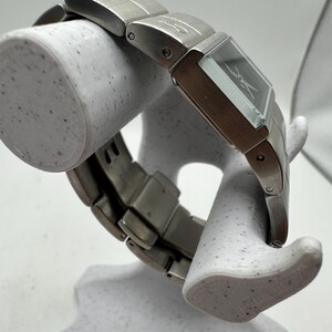 May include: A silver watch with a square face and a silver metal band. The watch is on a white display stand.
