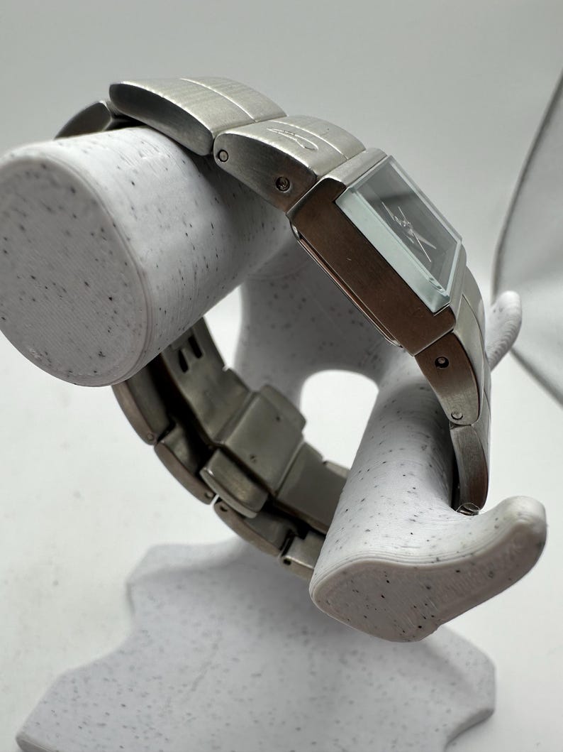 May include: A silver watch with a square face and a silver metal band. The watch is on a white display stand.
