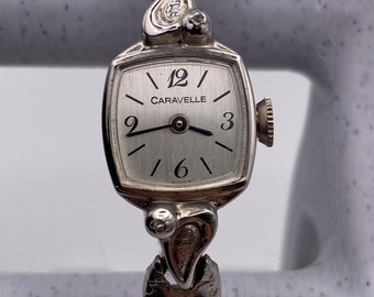 May include: A silver Caravel watch with a square face and a textured band. The watch face has black Roman numeral hour markers and a black minute hand. The watch has two small diamonds on the band.