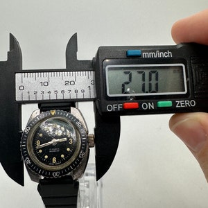 May include: A black and silver wristwatch with a black face and white markings. The watch has a black rubber strap and a rotating bezel. The watch is being measured with a digital caliper, which reads 27.0 mm.