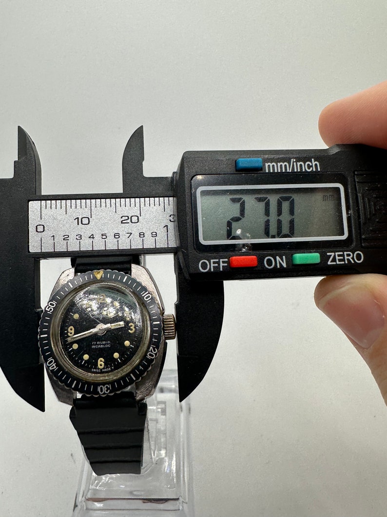 May include: A black and silver wristwatch with a black face and white markings. The watch has a black rubber strap and a rotating bezel. The watch is being measured with a digital caliper, which reads 27.0 mm.