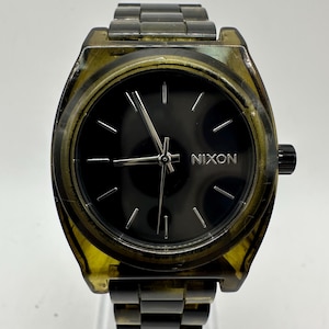 May include: A Nixon watch with a black face and silver hands. The watch has a transparent, amber-coloured case and a matching band. The watch face features the brand name "NIXON" in silver lettering.