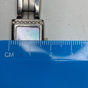 K&ouml;nnte beinhalten: Silberfarbene Uhr mit rechteckigem Zifferblatt und Perlmutt-Zifferblatt. Die Uhr hat ein silbernes Armband und ist mit kleinen Diamanten um die L&uuml;nette verziert. Das Zifferblatt der Uhr tr&auml;gt die Aufschrift "ODEON". Die Uhr wird mit einem Lineal gemessen und zeigt, dass sie etwa 2 cm breit ist.