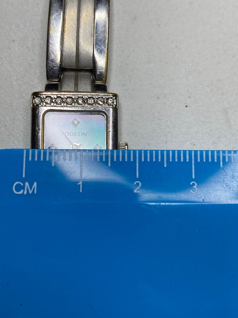 K&ouml;nnte beinhalten: Silberfarbene Uhr mit rechteckigem Zifferblatt und Perlmutt-Zifferblatt. Die Uhr hat ein silbernes Armband und ist mit kleinen Diamanten um die L&uuml;nette verziert. Das Zifferblatt der Uhr tr&auml;gt die Aufschrift "ODEON". Die Uhr wird mit einem Lineal gemessen und zeigt, dass sie etwa 2 cm breit ist.