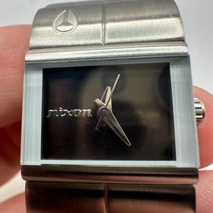May include: A silver Nixon watch with a black face and a silver band. The watch has a square face and a simple design.