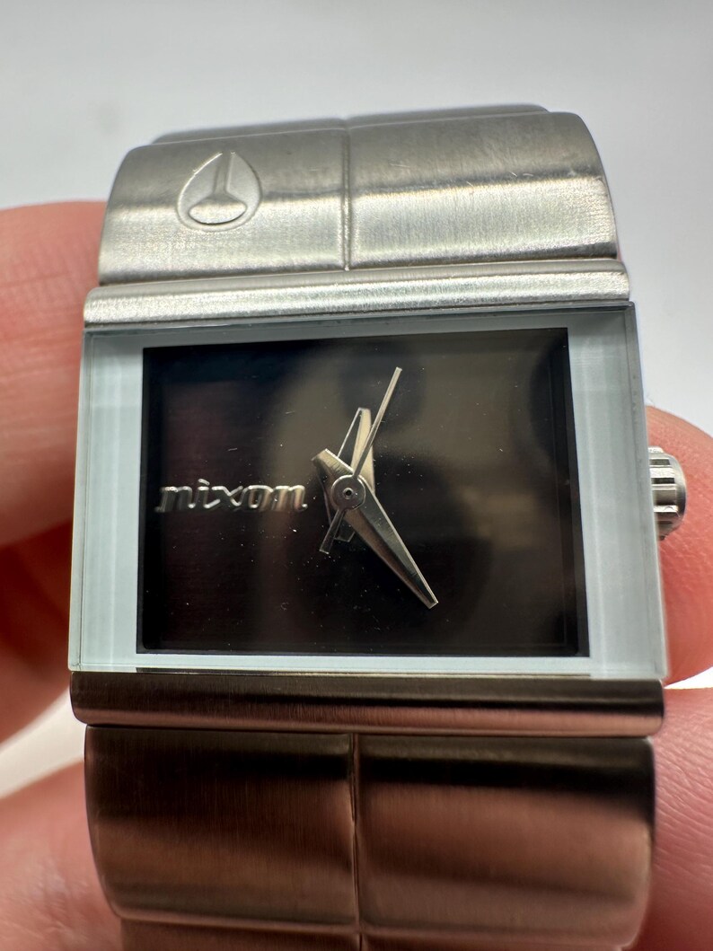 May include: A silver Nixon watch with a black face and a silver band. The watch has a square face and a simple design.