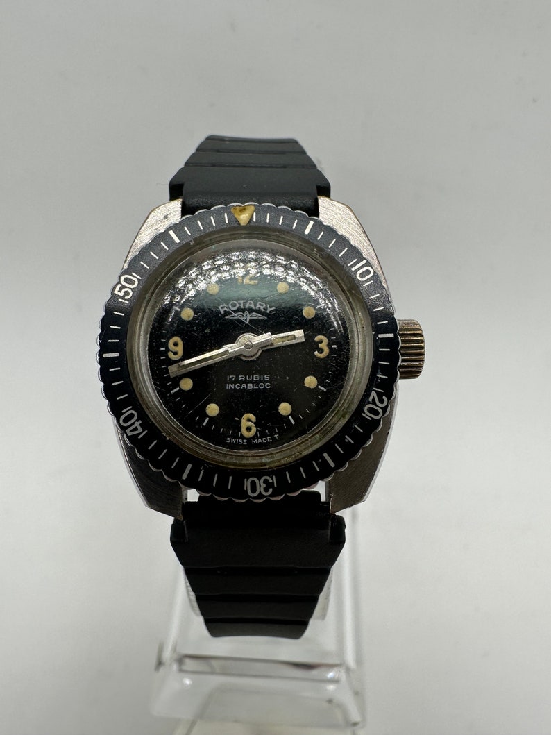 May include: A black Rotary wristwatch with a silver bezel and black rubber strap. The watch face has a black background with white markings and the text "Rotary", "17 Rubis", "Incabloc", and "Swiss Made T".