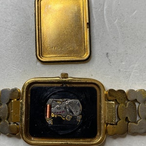 May include: A gold-toned rectangular wristwatch with the back cover removed, revealing the watch's inner workings. The watch band is a mix of gold and silver links. The back cover reads "SWISS CASE".