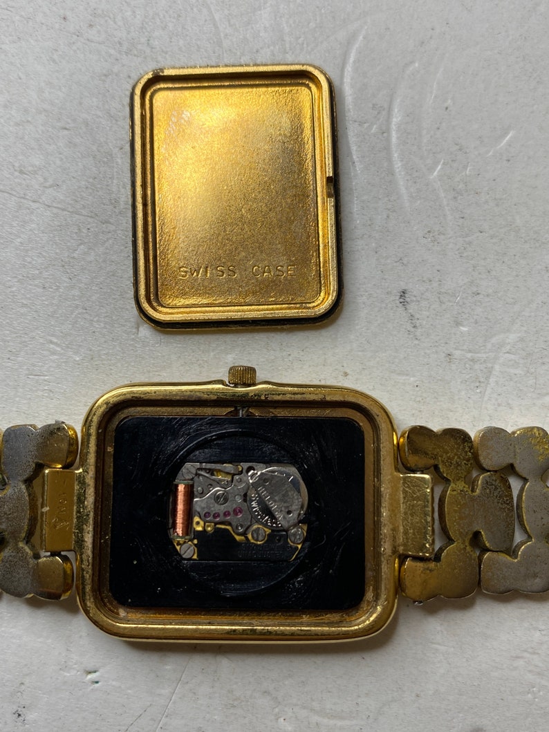 May include: A gold-toned rectangular wristwatch with the back cover removed, revealing the watch's inner workings. The watch band is a mix of gold and silver links. The back cover reads "SWISS CASE".