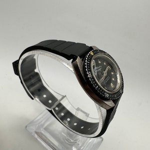 May include: A black wristwatch with a black rubber strap and a black bezel. The watch face is white with black markings and a black second hand. The watch has a date window at the 3 o'clock position.