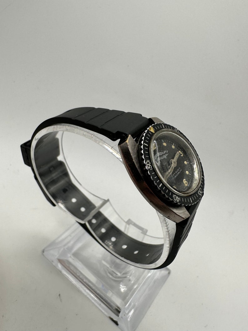 May include: A black wristwatch with a black rubber strap and a black bezel. The watch face is white with black markings and a black second hand. The watch has a date window at the 3 o'clock position.