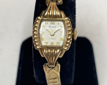 May include: A gold-toned wristwatch with a white face and black hands. The watch face has the words "Baron" and "17 Jewels" printed on it. The watch has a gold-toned metal band.