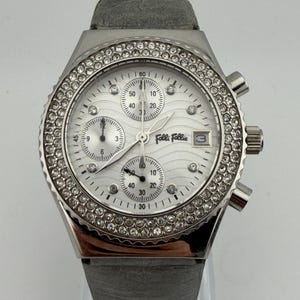 Può includere: Un orologio Folli Follie argentato con quadrante bianco e cinturino in pelle grigia. L'orologio presenta una lunetta incastonata con piccoli cristalli trasparenti. Il quadrante ha tre sotto-quadranti e un datario alle ore 3.
