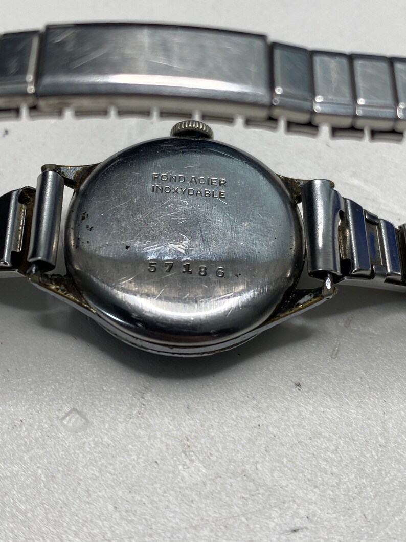 May include: Close-up of the back of a silver wristwatch with a stainless steel band. The watch face has the text "Fond Acier Inoxydable" and the number "57186".
