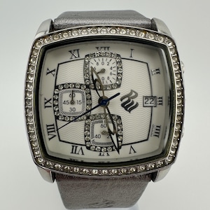 Può includere: Orologio da polso argentato con quadrante quadrato e cinturino in pelle grigia. Il quadrante bianco dell'orologio presenta numeri romani, tre quadranti secondari e un datario. La lunetta è incastonata con piccoli cristalli trasparenti.