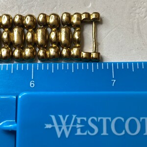 May include: Close-up of a gold-colored metal bracelet clasp. The clasp is made of connected oval and cylindrical links. A blue ruler with the word "WESTCOTT" is in the foreground, with measurements in inches.