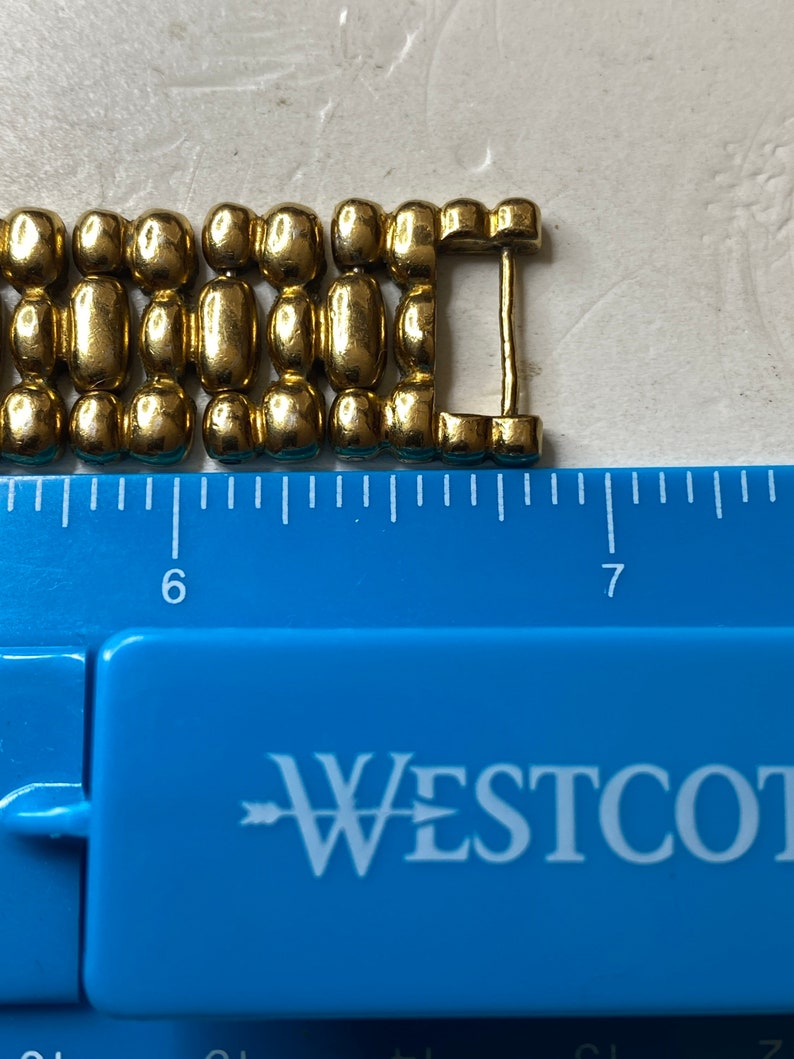 May include: Close-up of a gold-colored metal bracelet clasp. The clasp is made of connected oval and cylindrical links. A blue ruler with the word "WESTCOTT" is in the foreground, with measurements in inches.