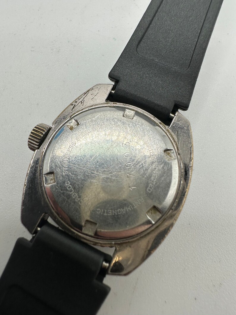 May include: The back of a silver wristwatch with a black rubber strap. The watch face has the text "Antimagnetic" and "25 Jewels" printed on it.