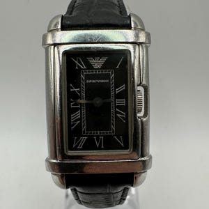 Puede incluir: Un reloj de pulsera rectangular plateado y negro con una correa de cuero negro. La esfera del reloj tiene marcadores de hora con números romanos y el nombre de la marca "EMPORIO ARMANI" está impreso en la esfera.