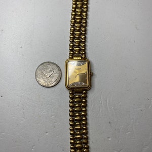 May include: Gold-toned wristwatch with a rectangular face and a beaded band. The watch face has a gold background with small, sparkling embellishments. The band is made of connected, rounded beads. The watch is next to a quarter.