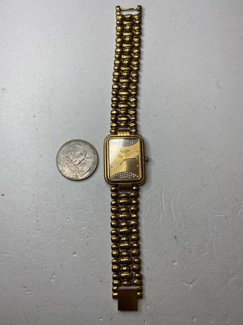 May include: Gold-toned wristwatch with a rectangular face and a beaded band. The watch face has a gold background with small, sparkling embellishments. The band is made of connected, rounded beads. The watch is next to a quarter.